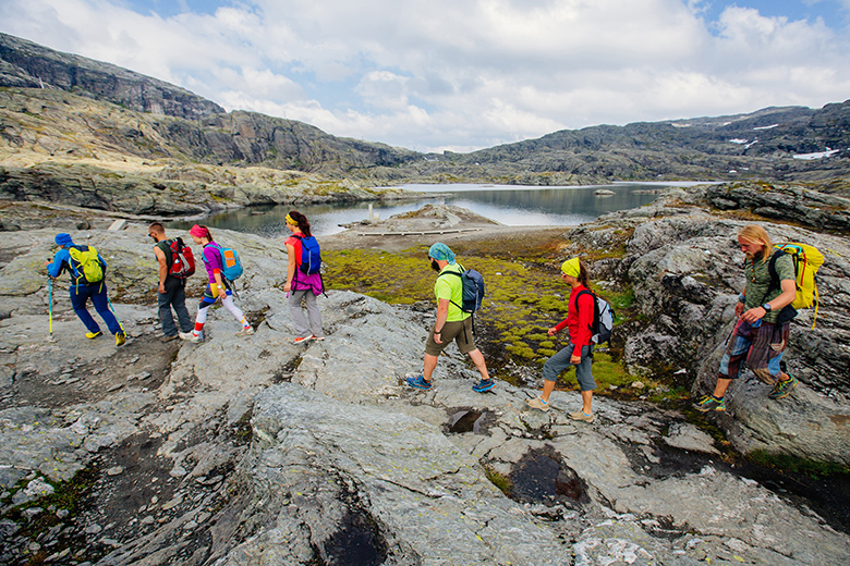 Group of seven hikers men and woman walking with backpacks and trekking poles along water lake in mountain valley during Norway travel.